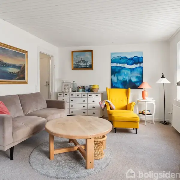 Gråt sofaarrangement med et rundt træbord står afslappet i en lys stue. Til højre er en gul lænestol, flankeret af moderne kunstplakater på væggen. En smal bogreol og dekorationer kompletterer rummet.