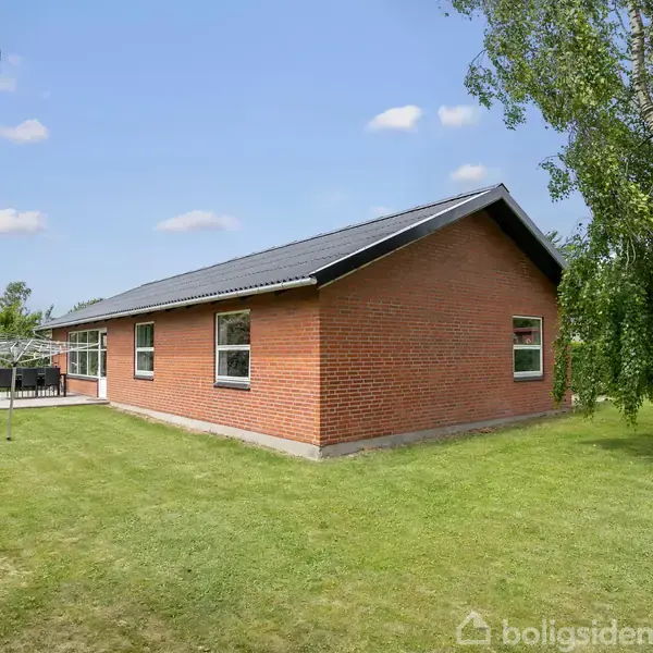 Et rødt murstenshus står stille i en velplejet have med græsplæne og træer. Huset har et sort sadeltag, og der ses en terrasse med havemøbler til venstre.