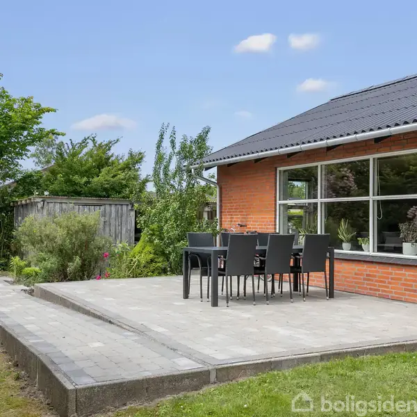 Terrasse med spisebord og stole, placeret foran et rødt murstenshus. Omgivet af grønne buske og træer under en blå himmel med spredte skyer.