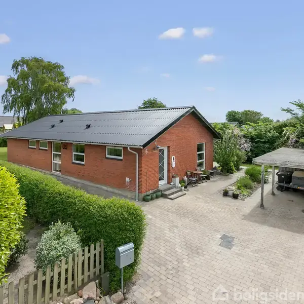 Rødt murstenshus med sort tag står stille i en have omgivet af grønne buske. En carport med en bil ved siden af en brostensbelagt indkørsel ses i forgrunden.