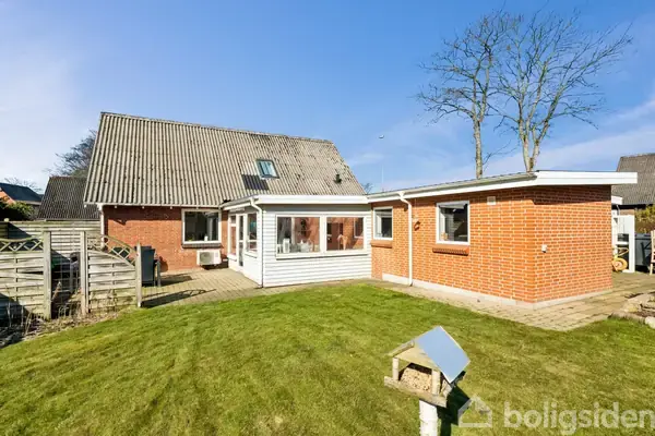 Rødt murstenshus med tilhørende udestue står i en have med velplejet græsplæne og et fuglehus. Omkring huset ses et plankeværk og et træ. Klar himmel i baggrunden.