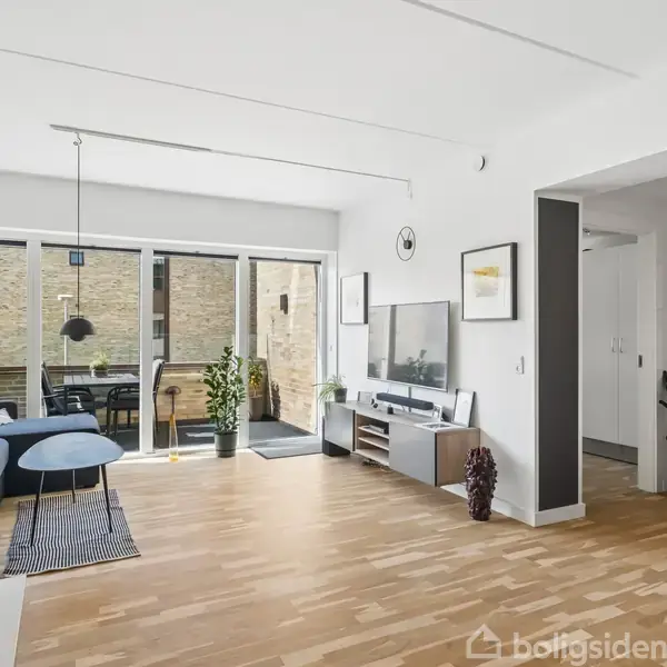 Stue med blå sofa og sofabord, vendt mod en stor glasdør til en balkon. TV-møbel mod højre væg. Lys trægulv og hvide vægge.