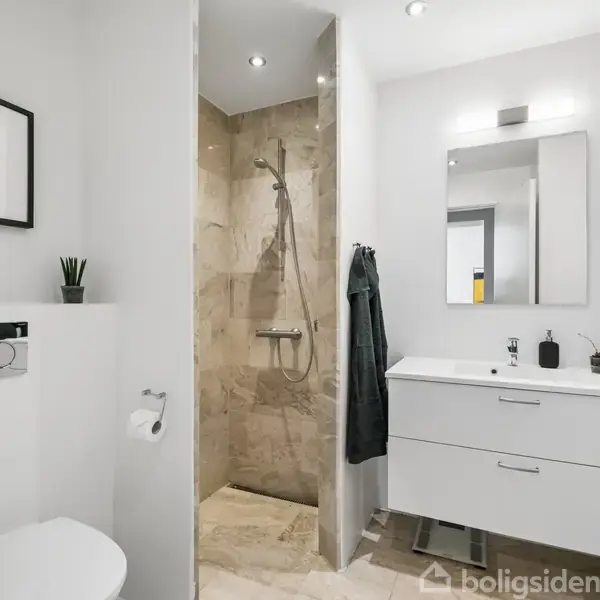 Badeværelse med hvidt inventar, viser toilet og bruser. Håndvask med skuffer og spejl, dekoreret med sæbedispenser og planter. Væggene er hvide med minimalistisk kunst.