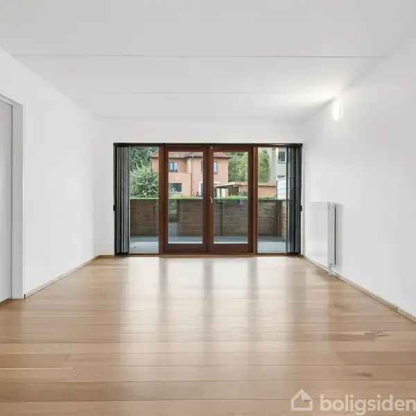 Et rummeligt, tomt værelse med trægulv har en stor glasdør, der åbner ud til en balkon. Værelset har hvide vægge og radiator, med udsigt mod nogle træer og en bygning.