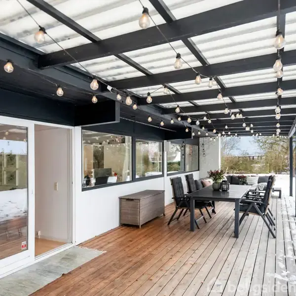 En overdækket terrasse med trægulv er dekoreret med hængende lyskæder. Der står et spisebord med stole. Sne dækker jorden udenfor, og glasdøre forbinder til huset.