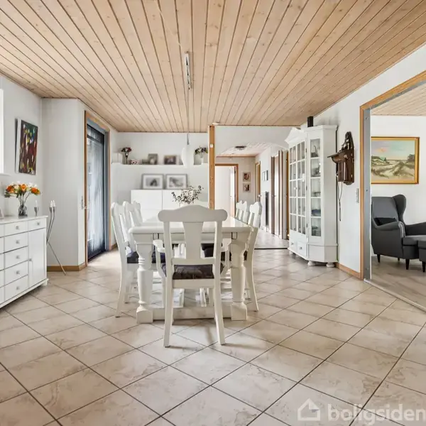 Et spisebord med hvide stole står centreret i et lyst køkken-alrum. Rummet har flisegulv, træloft og lyse møbler. Der er udsigt til en stue med sort sofa gennem en dør.