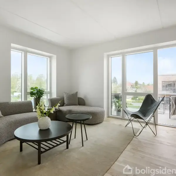 En moderne stue med grå sofa og runde borde, pyntet med en blomstervase. En dør fører til en balkon; store vinduer bader rummet i naturligt lys, omgivet af grønne omgivelser.