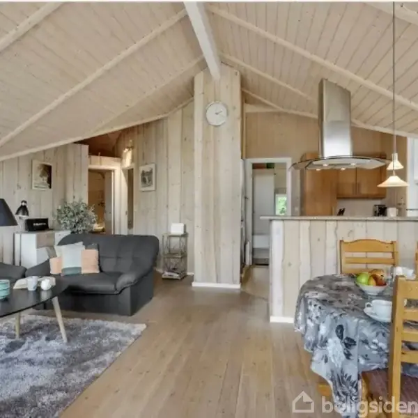 Stue med trævægge og højt loft indeholder en sort sofa, sofabord med dekorationer, spisebord med frugt og blomster, samt køkken i baggrunden. Tæppe på gulvet.