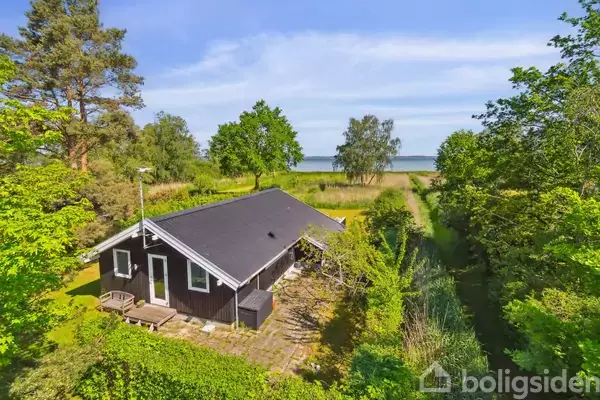 Et sort, træsommerhus med terrasse ligger omgivet af frodig vegetation, træer og buske. I baggrunden ses en sti, der fører til en strand med udsigt over vandet.