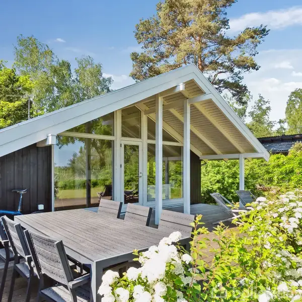 Et moderne sommerhus med glasfacade og skråt tag står i en frodig have. Udenfor er et stort terrassebord omgivet af stole, og en cykel læner sig op ad huset.