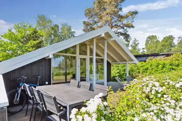Et moderne sommerhus med skrånende tag og store vinduer står i en grøn have. En træterrasse med havebord og stole er omkranset af blomstrende buske og cykel op ad huset.
