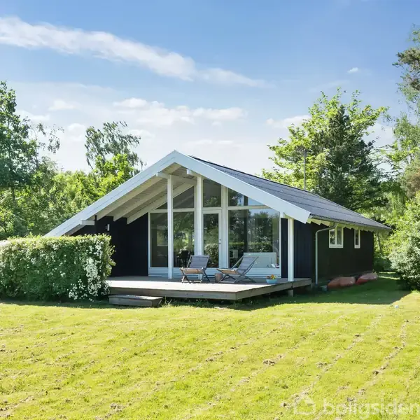 Sommerhus med sort træfacade står på en græsplæne; terrasse med to stole ses foran huset. Omgivet af træer og buske under en blå himmel.