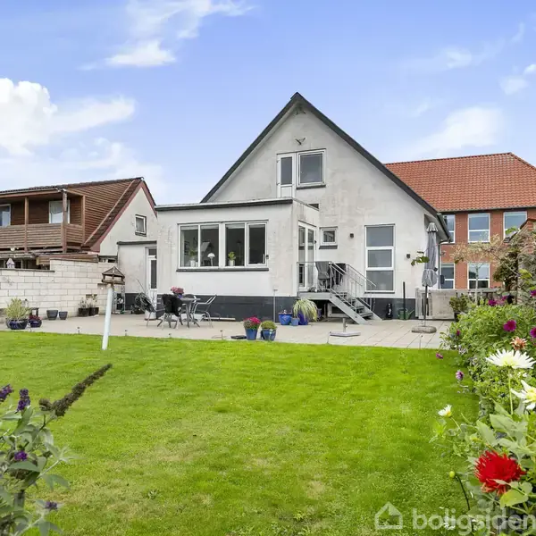 Huset står i en frodig have med grøn græsplæne og blomsterbede. Husets facade er hvid med rødt tegltag. Rundt om terrassen står havemøbler. Baggrunden viser flere nærliggende bygninger.