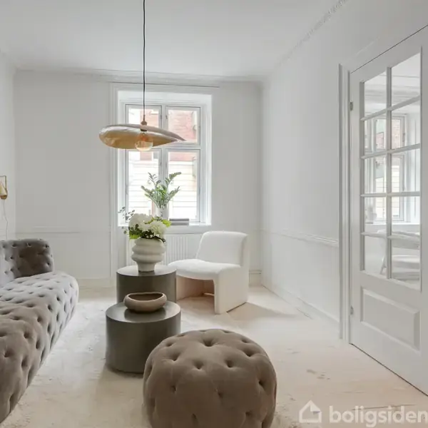 En grå, polstret sofa står i et lyst rum. To borde er foran sofaen, og en stor plante i hjørnet ved et vindue. En glasdør fører til et andet rum.