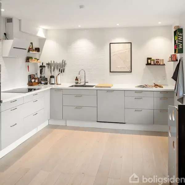 Lyst køkken med hvide skabe og træbordplade. En ovn og mikrobølgeovn er indbygget. Vindue til højre. Diverse køkkenredskaber og dekorative elementer pynter bordene. Ramme og "WINE"-kasse på væggen.