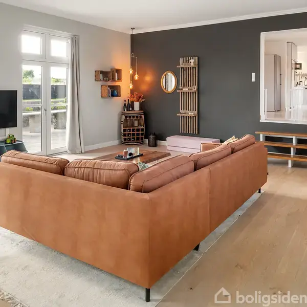 En brun sofa står i centrum af en lys stue med trægulve. En vægmonteret fladskærm og en reol ses nær vinduerne. Sort væg med kunst og runde spejle.