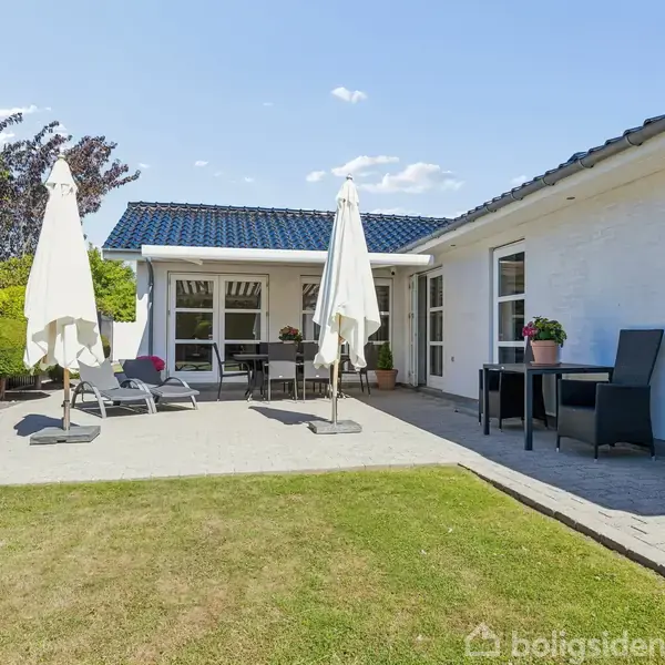Hvidt hus med blåttag, liggestole og haveparasol på terrasse, omgivet af grønne buske og græsplæne under blå himmel. Et bord og planter står ved vinduerne.