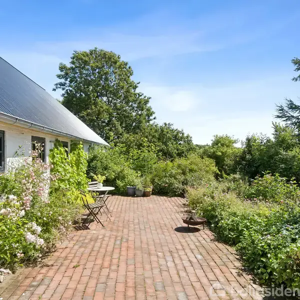 Hus med hvid facade og skråt tag. En terrasse med mursten og havemøbler til venstre. Omgivet af frodig vegetation og træer under en blå himmel.