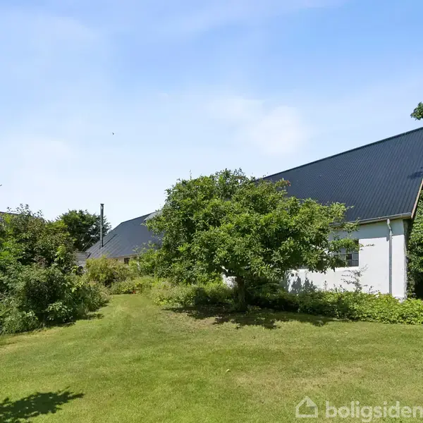 Hus med sort skråt tag står stille midt i en frodig have, omgivet af grønne træer og buske under en blå himmel.