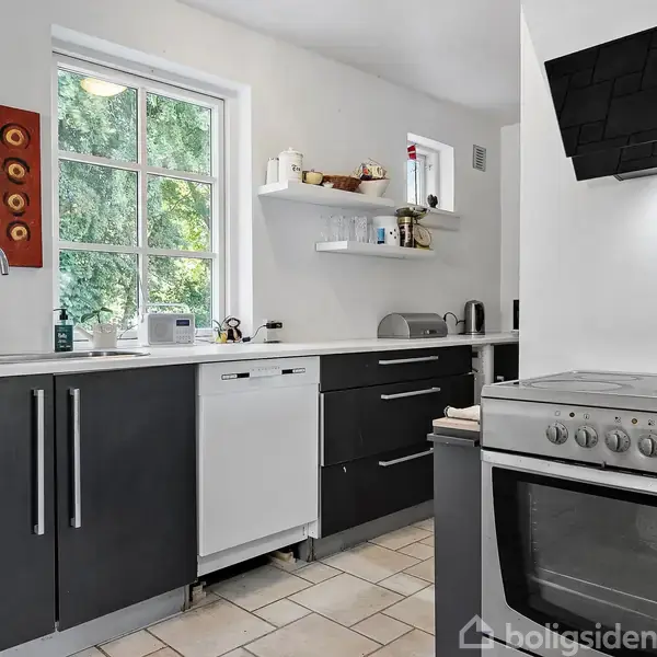 Komfur og ovn står ved siden af sorte køkkenskabe og en opvaskemaskine. Vindue med udsigt til grønt, vægbillede med farverige mønstre og hvide hylder i et lyst køkken.