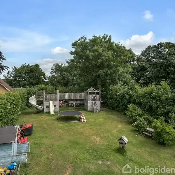 Et legeområde med et legetårn og rutsjebane i en grøn have. Der er også en trampolin og forskellige legeredskaber, omgivet af buske og træer under en blå himmel.