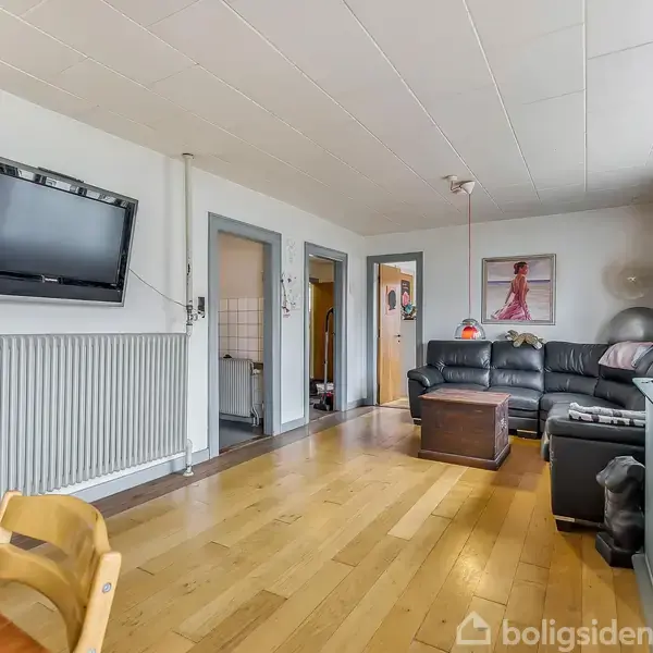 Fjernsyn hænger på en væg over en radiator i en stue med trægulv. Til højre står en sort lædersofa ved siden af et stort vindue med lysindfald.