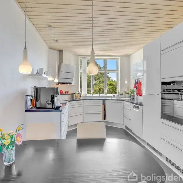 Køkkenbord med vase med blomster står i et hvidt moderne køkken med indbyggede apparater. Store vinduer slipper naturligt lys ind, mens lamper hænger ned fra træloftet.