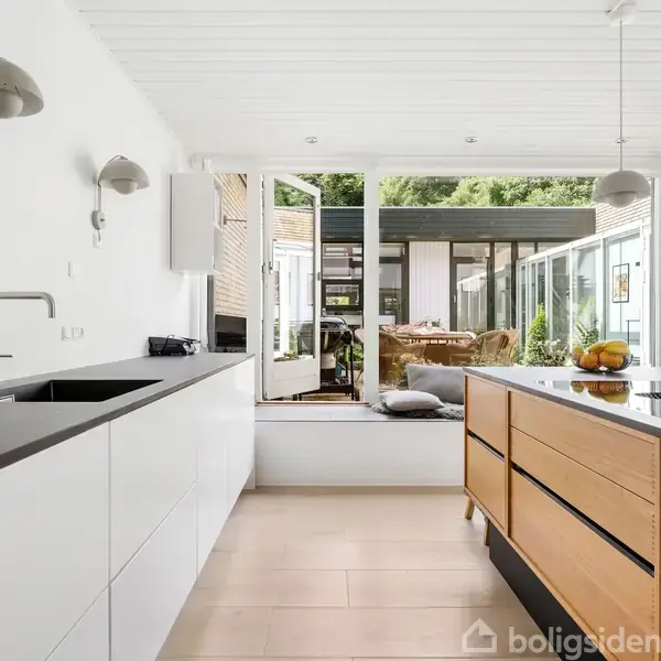 Et moderne køkken med sorte og træfarvede elementer, ser ud mod en lys havestue med åbne døre. Udenfor er der grønt.