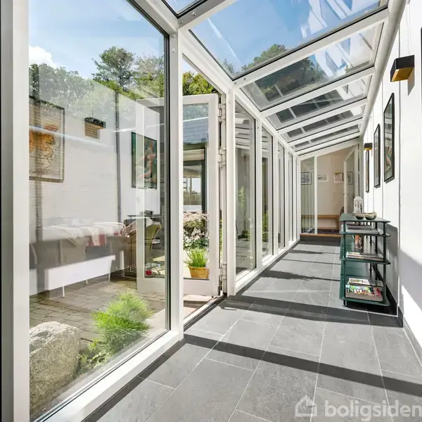 En glasbeklædt gang fyldt med lys. Handling: Lyset strømmer ind udefra. Kontekst: Omgivet af minimalistiske møbler og planter i et moderne hjem.