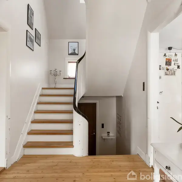 Trappeopgang med hvidmalede vægge og trætrin, dekoreret med indrammede billeder. Til venstre er en stue med grå sofa, og til højre et skab med spejl og plante.