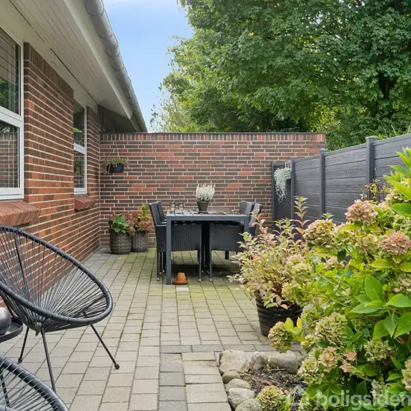 Et par stole står på en flisebelagt terrasse ved et murstenshus. En sort bordgruppe med planter i krukker tilføjer atmosfære, omgivet af farverige buske og et hegn.