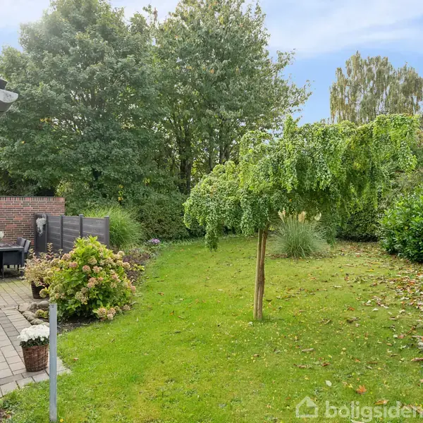 En grøn have med en hængepil i midten, omgivet af buske og træer. En terrasse til venstre med et bord og stole foran en murstensbygning.
