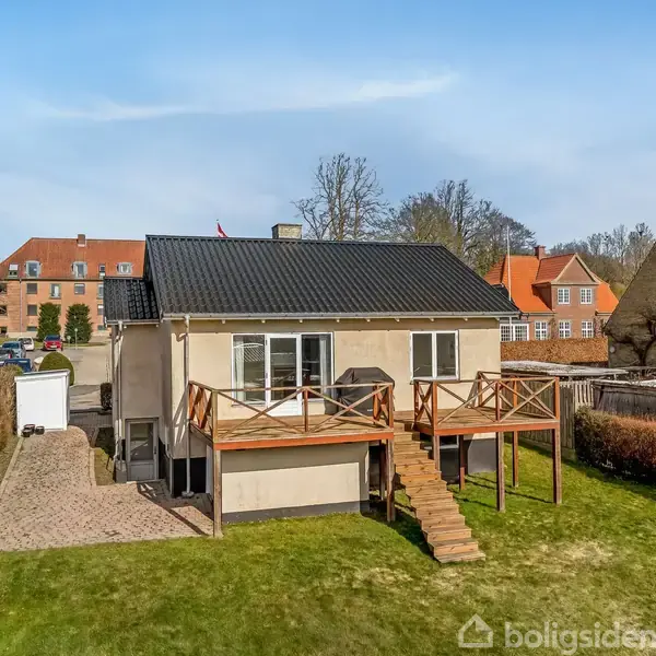 Huset, placeret i en have, har en trappe førende til en hævet terrasse. Kontekst: nabolag med flere huse og en brolagt indkørsel langs venstre side.
