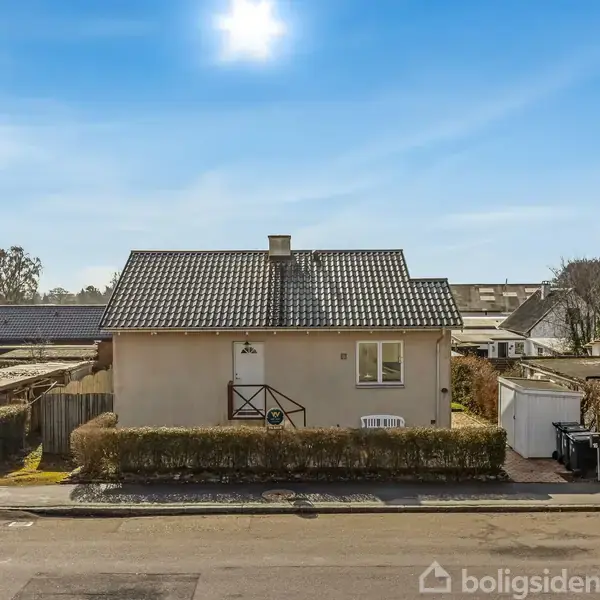 Et lille, hvidt hus med gråt tag står stille på en stille vej. Hækken omkranser grunden, og solen skinner fra en blå himmel.