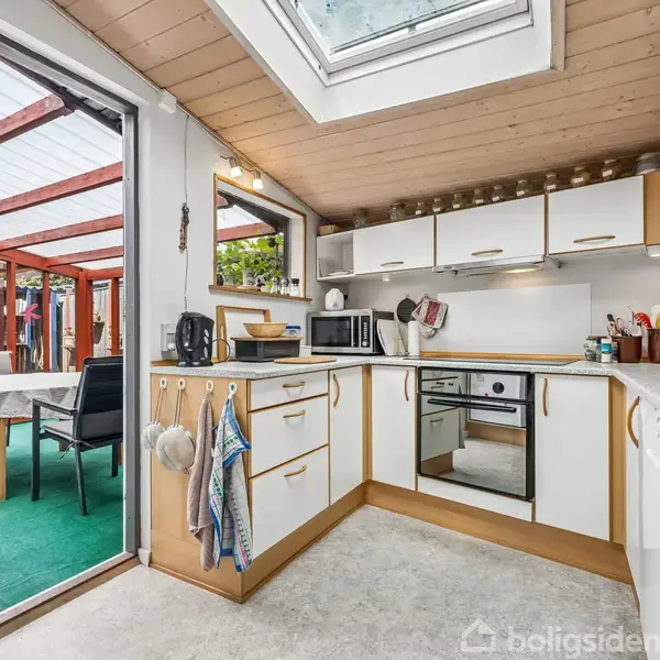 Køkken med hvide skabe og moderne apparater, som åbner ud mod en overdækket terrasse med grøn tæppebelægning og spisebord, skaber en lys og indbydende atmosfære med ovenlysvindue.