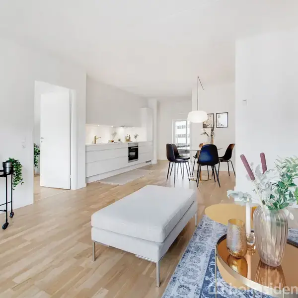 Stueområde med sofa, sofabord og plante; udsigt til moderne køkken med spisebord til højre; trægulve og lys indretning. Atmosfæren er lys og moderne med minimalistisk design.