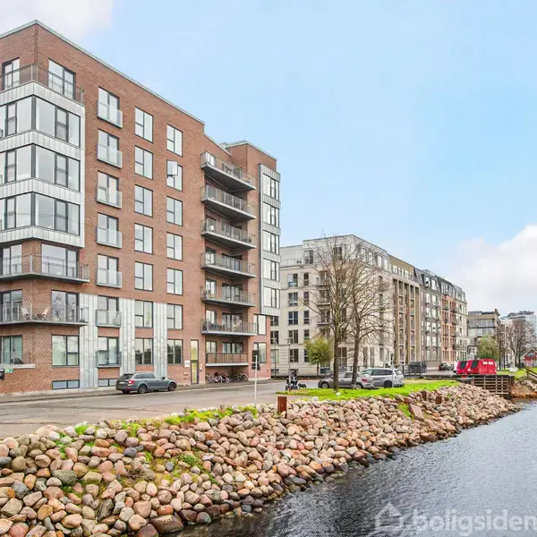 En moderne lejlighedsbygning står ved en flod med en stenet bred. Flere biler er parkeret langs vejen foran bygningen, og andre boligkomplekser ses i baggrunden.