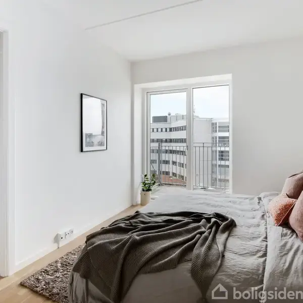 Soveværelse med dobbeltseng dækket af tæpper og puder. Store vinduer åbner mod en balkon med udsigt over bygningsfællesskab. Et billede hænger på den hvide væg ved siden af en plante.