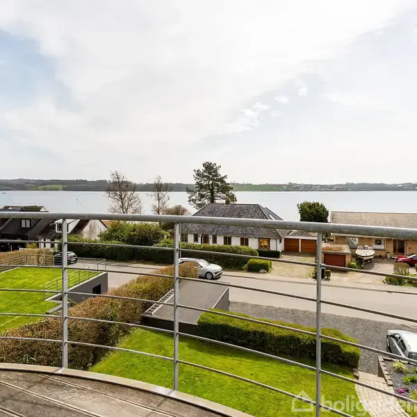 Balkon med metalgelænder, der vender mod parcelhuse og en vej. I baggrunden ses en bred udsigt over en rolig sø eller fjord og fjerne bakker.