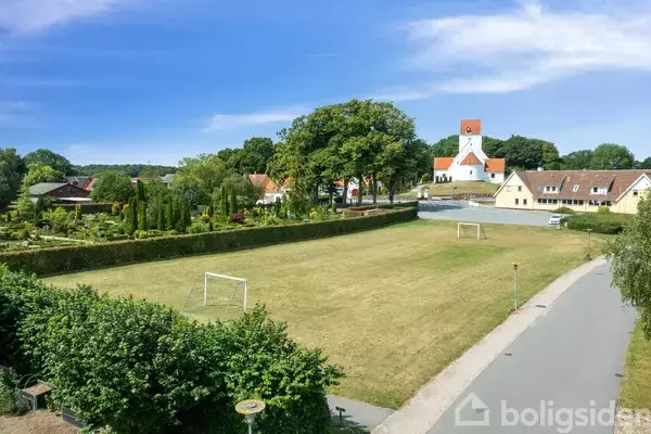 En fodboldbane med mål står ubenyttet. Den er omgivet af hække og en sti til højre. I baggrunden ses en kirke og flere huse.