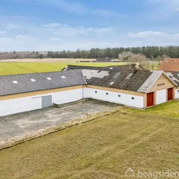 Stor landbrugsbygning med delvist ødelagt tag er omgivet af marker. Bygningen er hvid med røde porte, beliggende i en landlig, skovpræget region.