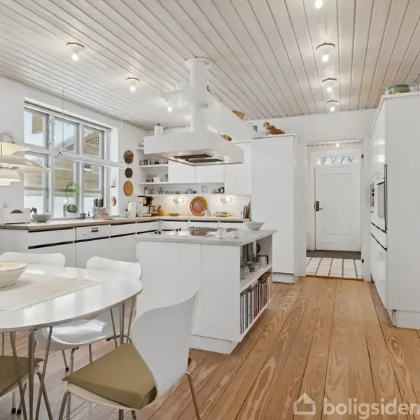 Hvidt køkken med trægulv og spisebord. Bordet har fire stole og en lampe hænger over det. Skabene og apparaterne er moderne, og store vinduer giver naturligt lys.