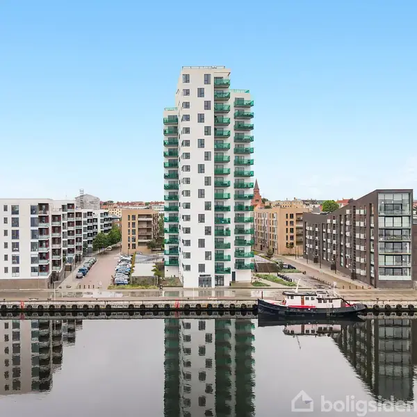 En høj, moderne bygning med mange etager står ved en kaj. Bygningen er omgivet af vand og andre lejlighedskomplekser i et byområde under en klar blå himmel.