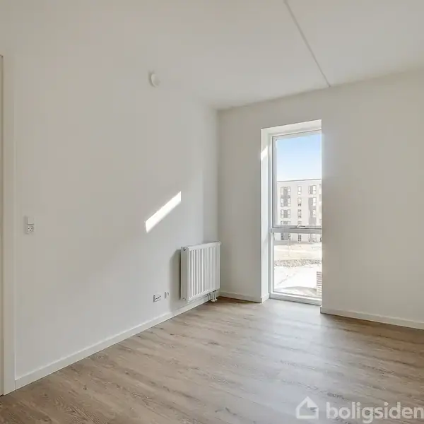 Et lyst rum med trælaminatgulv har en stor, lys fyldt vinduesdør til højre. Til venstre fører en åbning til et tilstødende værelse med indbyggede skabe.
