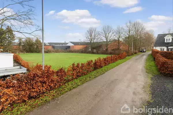 En græsplæne strækker sig bredt, kantet af en række buske, mens træer og bygninger ses i baggrunden under en lysende blå himmel.