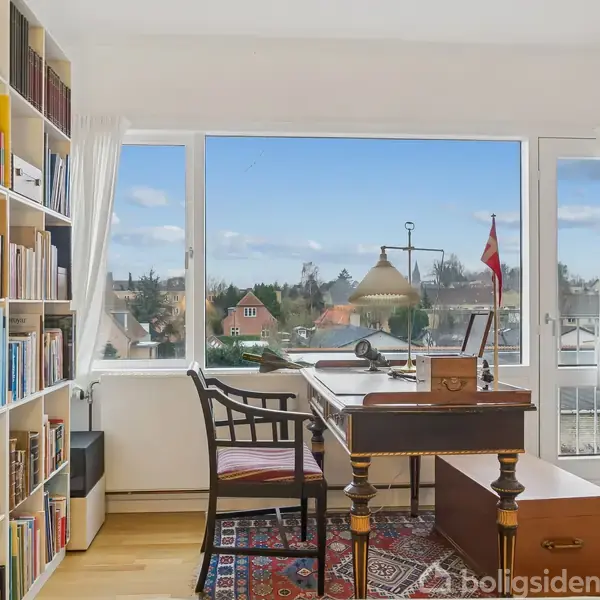 Et skrivebord med en lampe og bøger står foran et stort vindue med udsigt over huse og flag. Bogreol til venstre, tæppe på gulvet, lys og luftig atmosfære.
