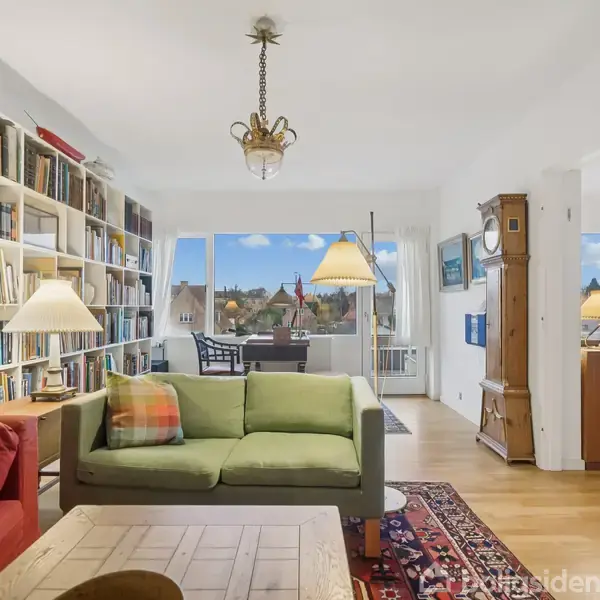 Stue med grøn sofa og rødt tæppe, omgivet af fyldte bogreoler. Store vinduer åbner op til en byudsigt. Et spisebord med stole ses i baggrunden. Elegant belysning.