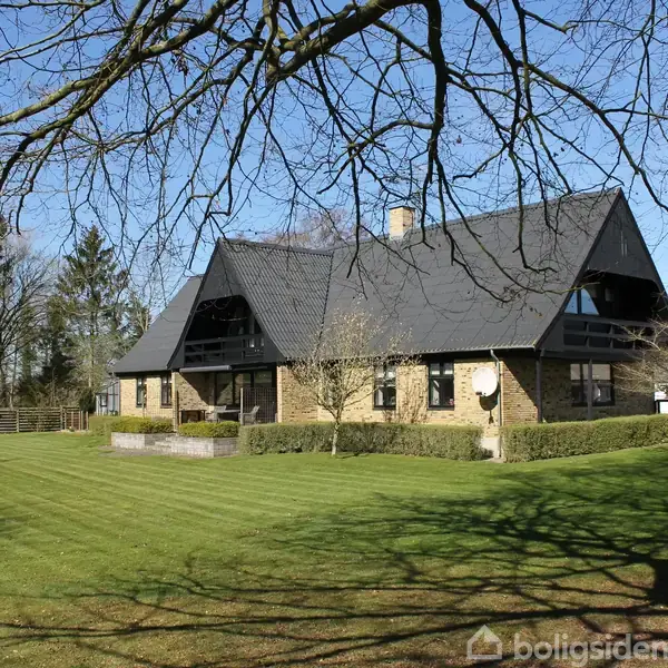 En stor moderne villa med stejl taghældning står blandt træer, omkranset af en velplejet græsplæne i et åbent, grønt haveområde.