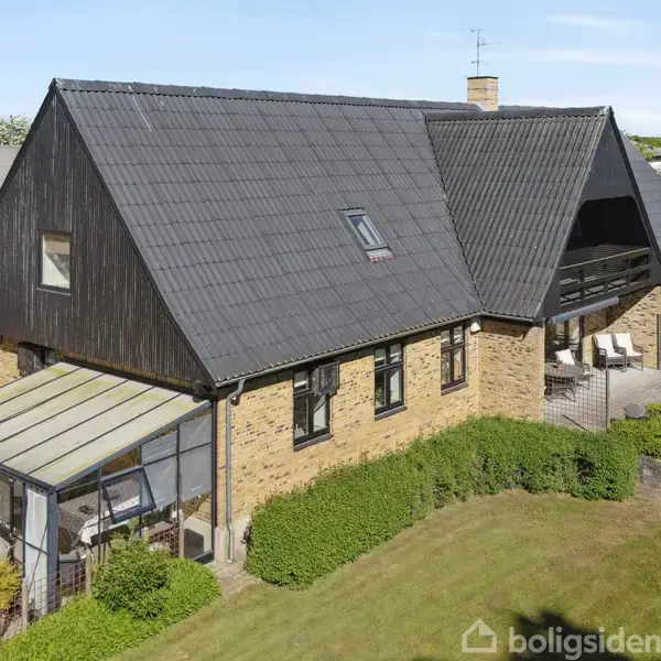 Et moderne boligudarhus med sort tag og gul væg står i en grøn have. Tilbygning af glas ser ud som en udestue, omgivet af buske og plæne.