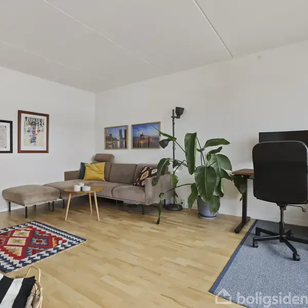 En stue med en sofa og et sofabord står ved siden af en arbejdsstation med en skærm og kontorstol. Væggene har kunst, og en stor plante står i hjørnet.